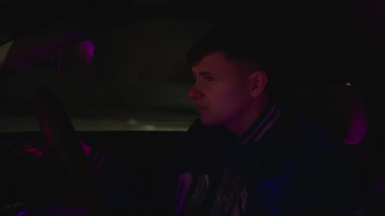 Auton sisätila yöllä, valkoihoinen kuljettaja mietteliäänä ajelemassa. Synkkä neonpurppura hohde, college-takki, vakaat kädet ratissa, kojelaudan valot välkkyvät, yksinäinen hidas ajomatka, siirtyminen roolista toiseen opiskelujen välissä