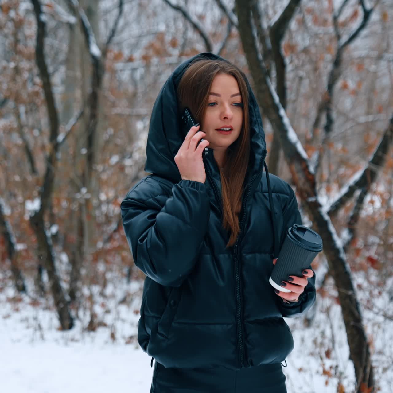 Long-haired brunette walking outdoors in winter. Girl wearing black jacket drinks coffee and chats on the phone