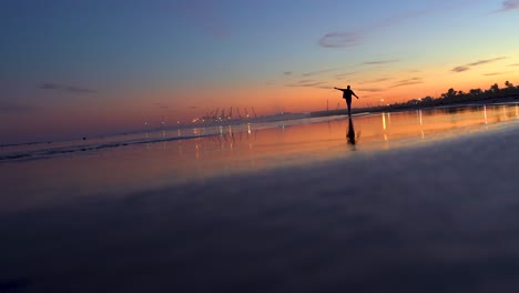 Man-turns-around-and-makes-the-plane-on-the-beach-with-arms-outstretched-at-sunset,-beautiful-backlight-with-the-sloping-beach,-the-sea-and-the-port-with-cranes-in-the-background