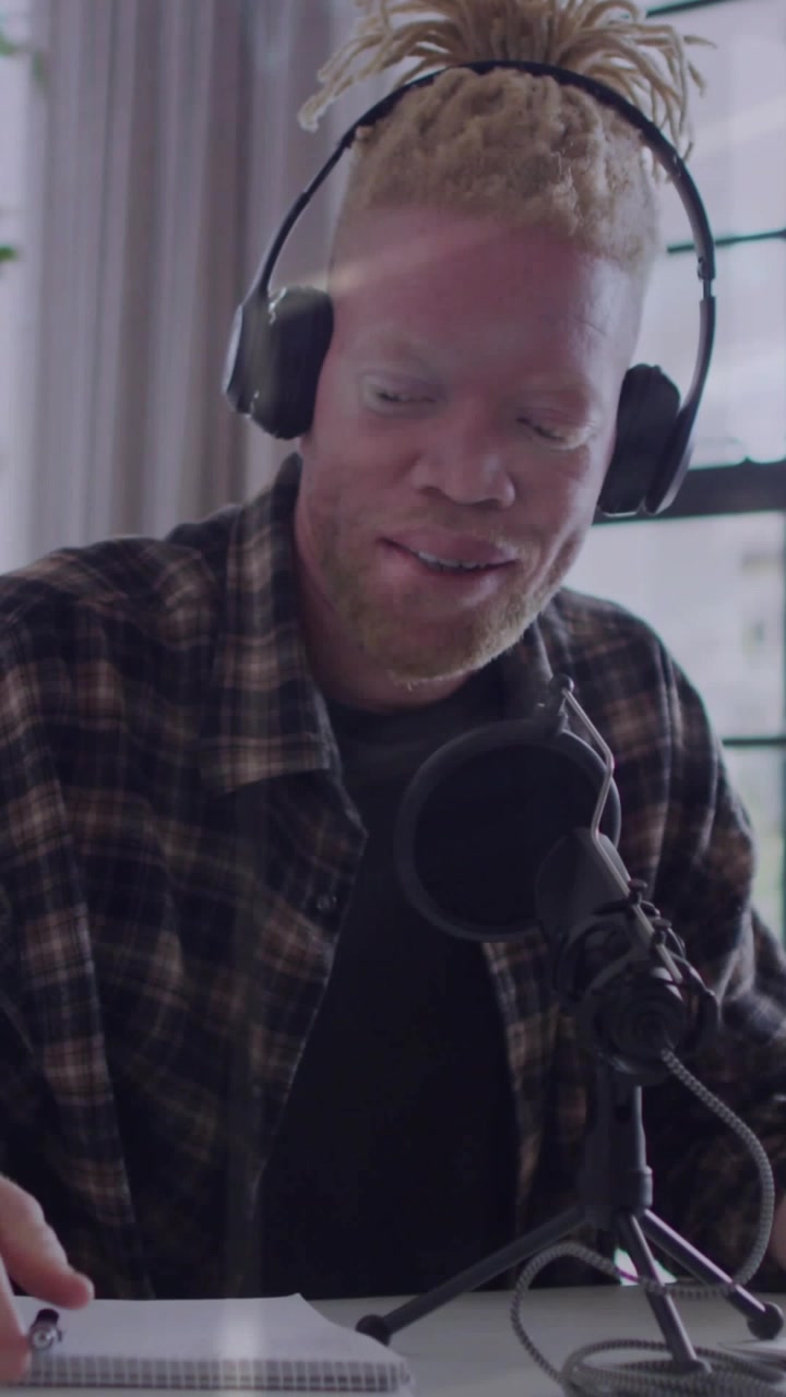Vertikal video: Podcaster justerer mikrofon og noter, optager, taler, smiler med genskin fra vinduet.