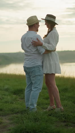 dos personas felices se abrazan mientras bailan en una colina cubierta de hierba al lado de un lago tranquilo. el hombre está vestido con una camisa blanca, sombrero y vaqueros, mientras que la mujer lleva un vestido blanco y sombrero negro