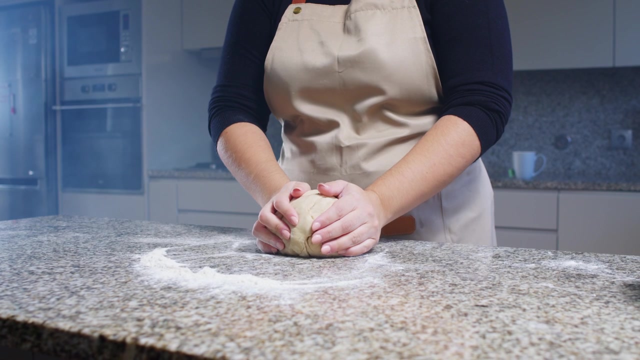A woman shapes dough on a flour-dusted countertop, preparing it for baking. Cinematic lighting enhances the kitchen scene as she works the dough.