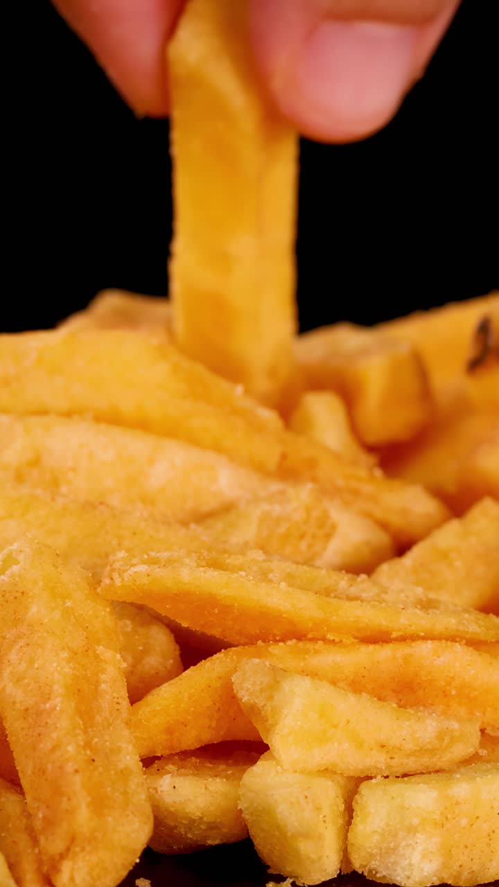 Mão seleciona uma única batata frita crocante de uma pilha sob luz brilhante, fundo preto, visão macro.