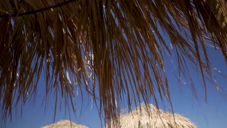 Toma-Manual-De-Hojas-De-Palma-Secas-De-Un-Paraguas-Tiki-Hut-Moviéndose-Por-El-Viento-Que-Sopla-En-Un-Día-Soleado-Y-Ventoso-En-Santorini,-Grecia