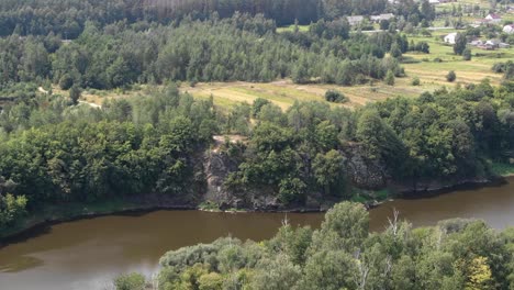 Aerial-View-of-River-Bordered-by-Forest-Approaching-Cliff-With-Field-in-Background