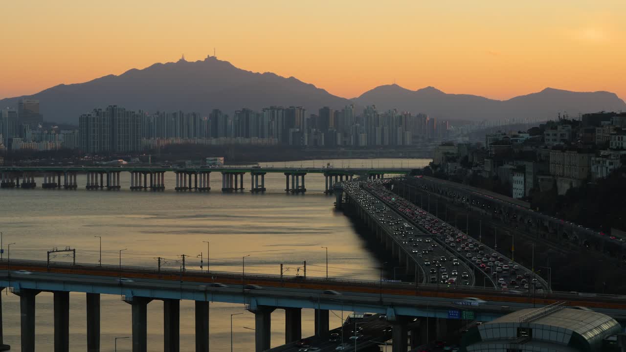 Timelapse del paesaggio urbano di Seul al tramonto con intenso traffico sul ponte Donghodae e sull'autostrada Gangbyeonbuk-ro, montagne panoramiche in silhouette sullo sfondo di un cielo arancione brillante illuminato dal sole in inverno