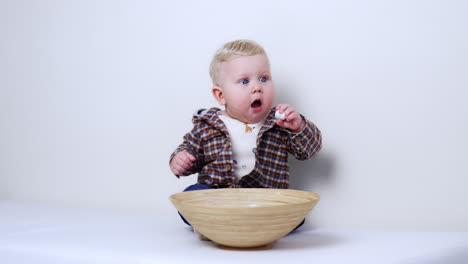 beautiful blond caucasian baby in checkered shirt sits at a big bowl. kid picks a marshmallow and pulls it to his mouth. white backdrop.