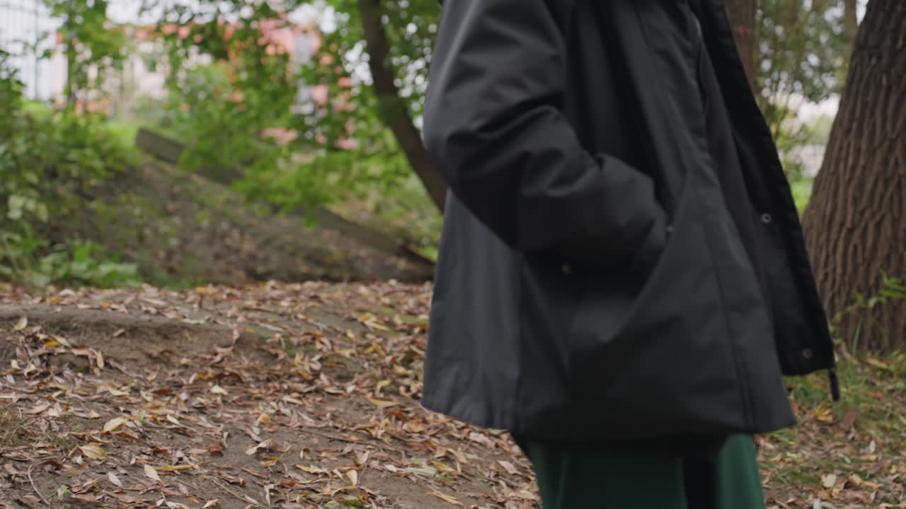 Woman walking on forest trail, pausing to sit and gaze upward with contemplative expression wearing dark jacket and scarf, sturdy sneakers on leafstrewn ground, soft overcast light, quiet autumn