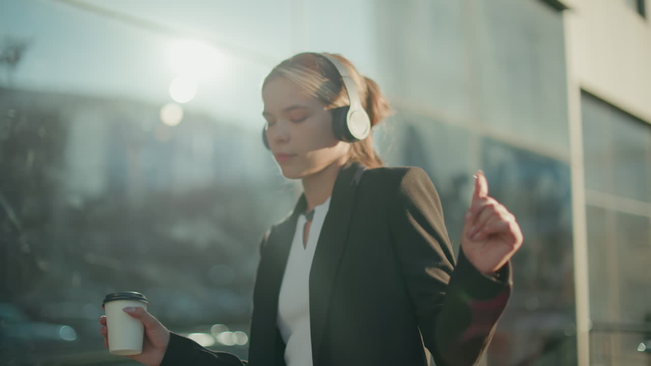 Young girl in headset dancing to music while holding takeaway coffee in urban setting, sunlight creating glow on her face with glass building reflecting surrounding environment in background
