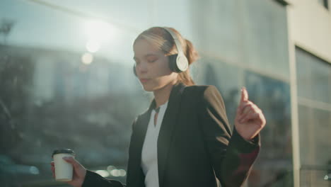 young girl in headset dancing to music while holding takeaway coffee in urban setting, sunlight creating glow on her face with glass building reflecting surrounding environment in background