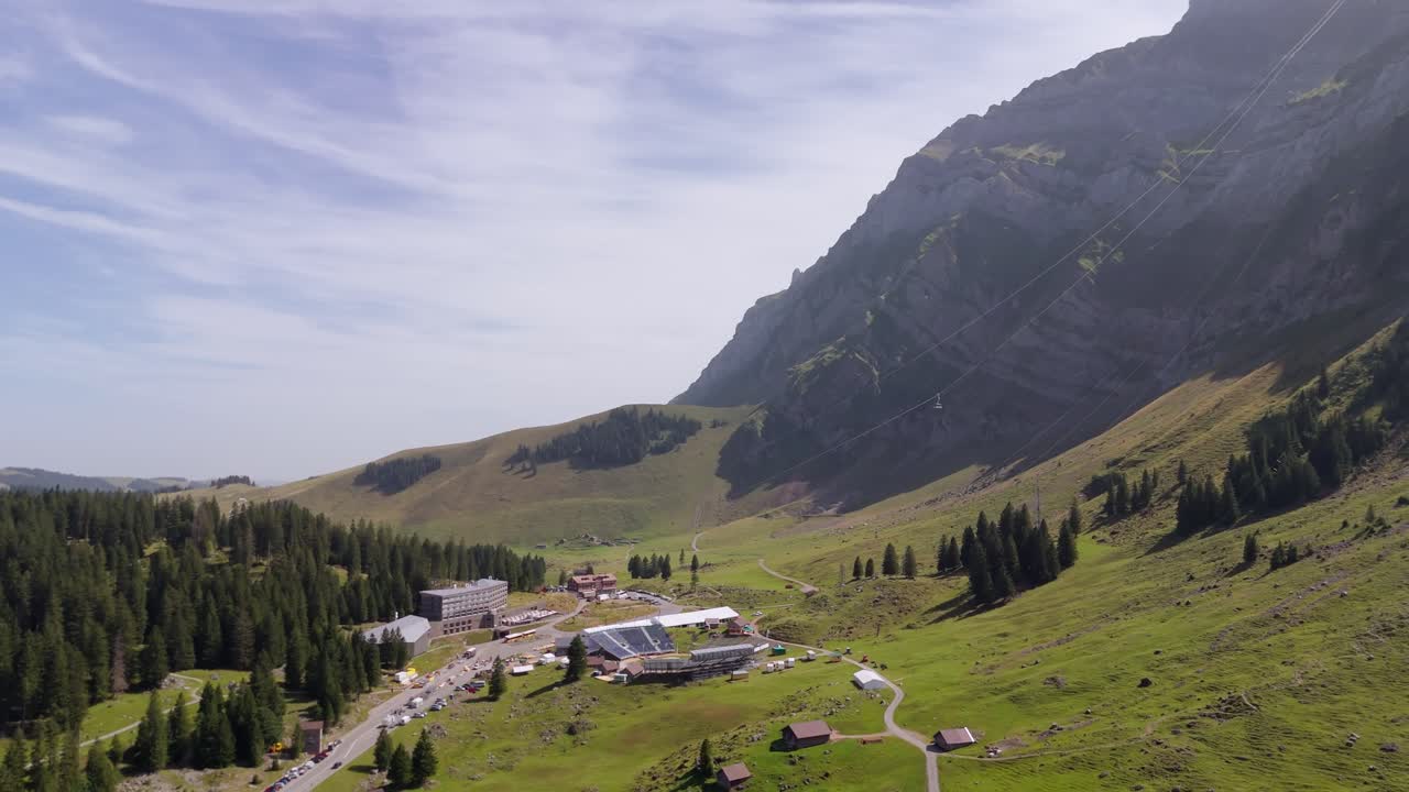 Forward drone over Schwagalp beneath Santis, showing a high altitude mountain hotel, cable car lines, alpine pastures and wooden shepherd huts at a Swiss ski resort in summer