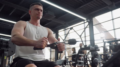 muscular man seated on bench pulling cable rowing machine with intense grimace showing effort and strength in gym with blurred people in background displaying focused fitness training form