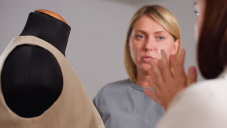 female designers evaluate beige waistcoat on black mannequin, fingertips tracing panel while exchanging comments, checking fabric tension, seam alignment, silhouette during focused studio session