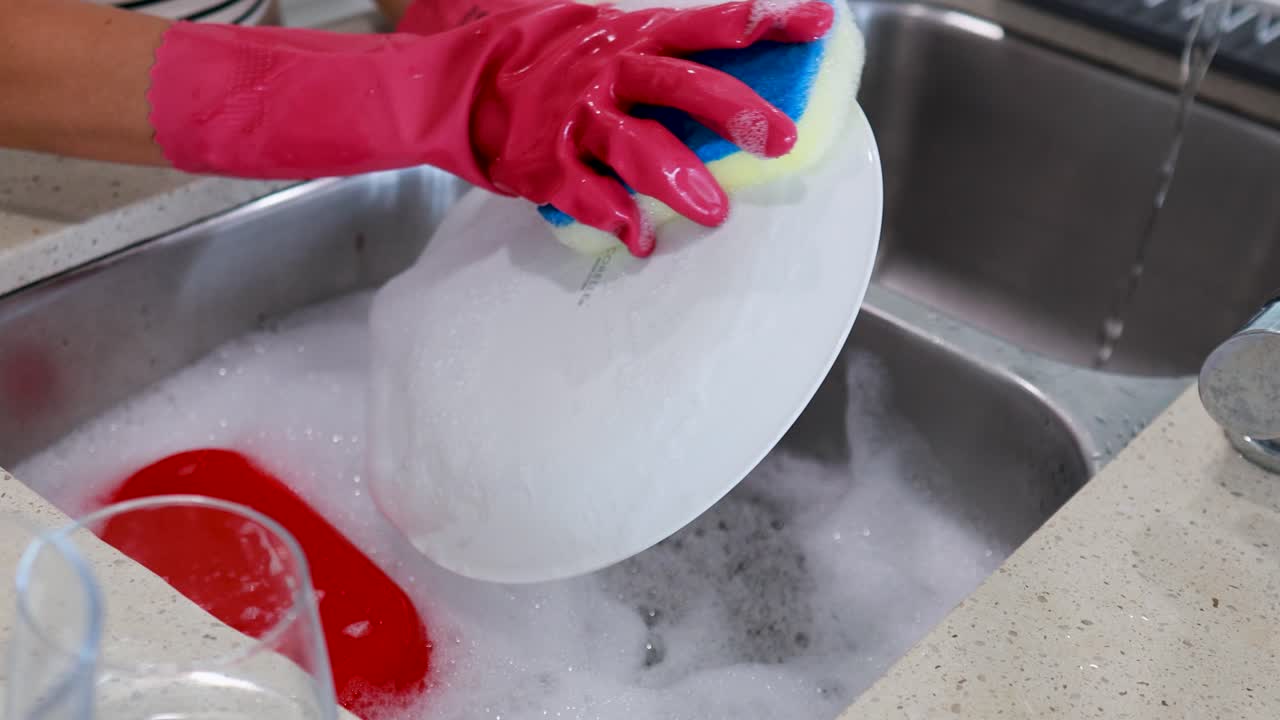 Hands in gloves wash dishes with a sponge in a kitchen sink. Bright lighting highlights the cleaning process