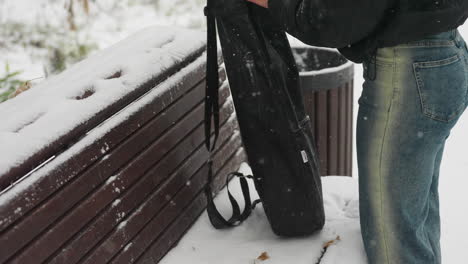 partial view of lady removing guitar pack to place on snow-covered bench in snowy park, with blurred figures walking in distant background surrounded by frosted trees