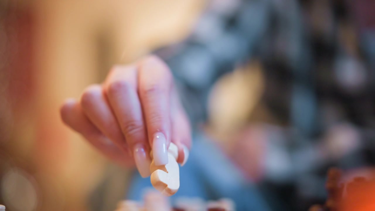Close up of hand moving chess pieces on board focusing primarily on white pawns and rook. background is softly blurred with warm lighting from orange candle creating intimate indoor ambiance