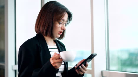 brunette lady is focused on the her smartphone. woman in glasses stands at the window drinking coffee and talking.