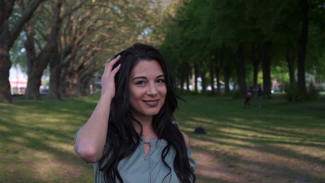 mujer latina atractiva y juguetona con cabello negro ondulado caminando bajo los árboles en un parque en londres, mirando a la cámara, feliz con una hermosa sonrisa