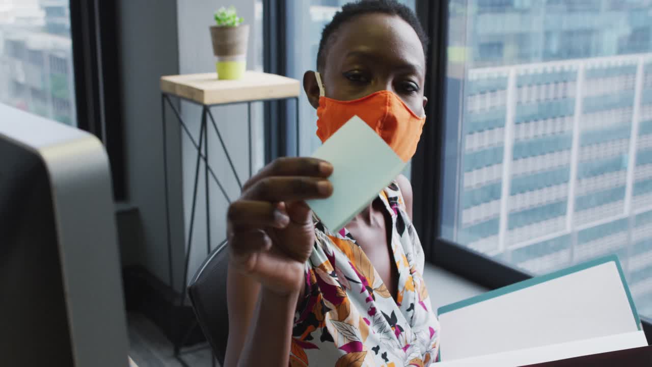 Donna afroamericana con la mascherina che legge un appunto seduta alla sua scrivania in un ufficio moderno.