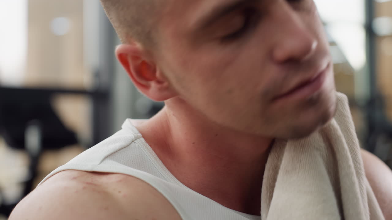 Athlete wipes face and neck with towel after intense gym workout under natural light showing sweat glistening skin texture and closeup hand motion in modern fitness center environment
