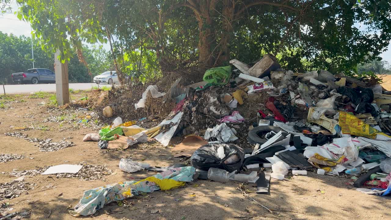 Foto estática e ampla de uma grande pilha de lixo plástico descartada sob uma árvore à beira da estrada