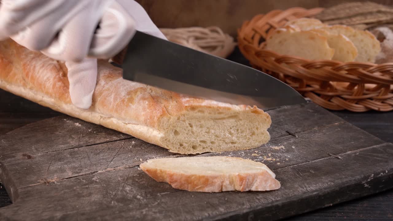 The chef cuts fresh crispy baguette into slices with kitchen knife. Close-up