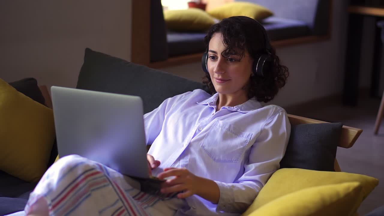 mujer elegante, morena trabajando en una computadora portátil en la oficina de casa. mujer joven sentada cómodamente en un sofá y navegando. trabajo independiente remoto o estudiando - con auriculares, sonriendo. interior moderno