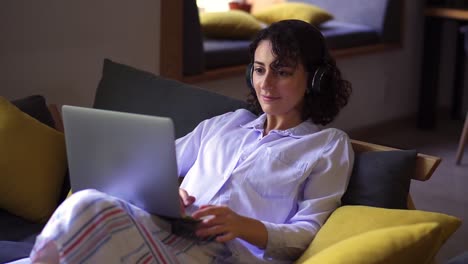 stylish, brunette woman working on laptop at home office. young female sitting comfortably on a couch and browsing. remote freelance work or studying - in headphones, smiling. modern interior