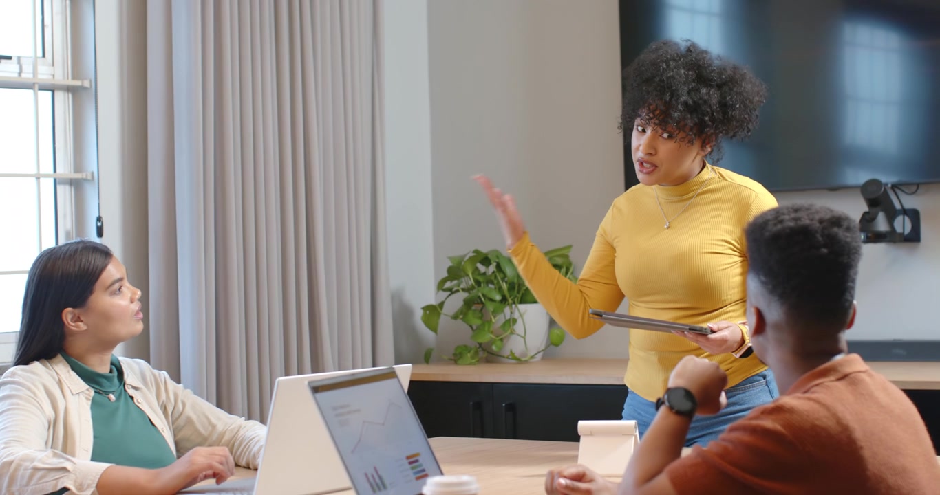 Diverse team watching African American woman using tablet, explaining data at table with laptops