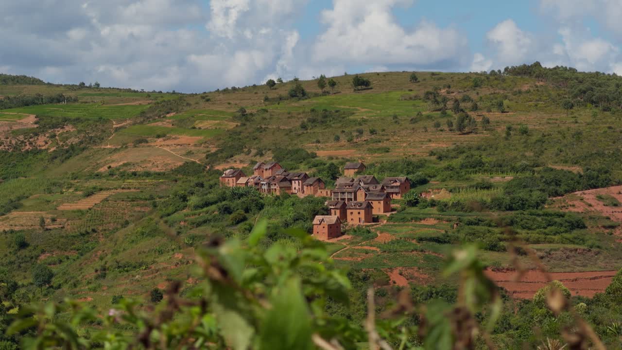 Un'ampia inquadratura panoramica che mostra un villaggio tradizionale con case in mattoni annidate su un lussureggiante pendio verde sotto un cielo azzurro nuvoloso negli altopiani rurali del Madagascar. AfricaVillage, Madagascar, Altopiani.