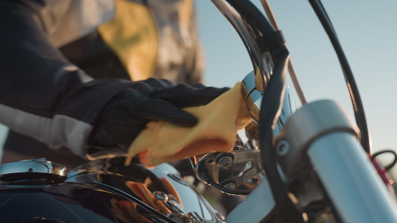 Man in leather jacket wearing gloves wipes motorcycle handle with yellow cloth, focusing on chrome details and black finish, outdoor maintenance scene with blurred background under bright daylight