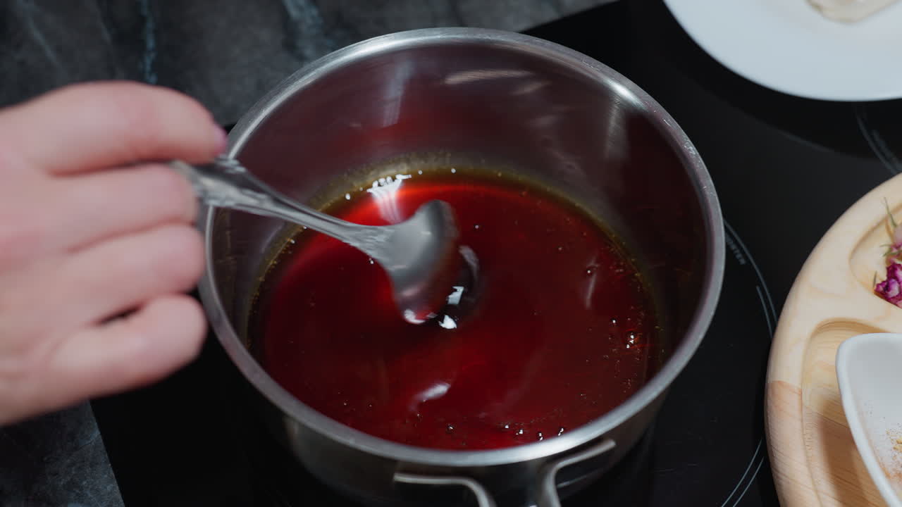 primer plano de una persona blanca moviendo a mano aceite caliente en una olla de acero inoxidable usando una cuchara en una estufa eléctrica, con una vista parcial de la bandeja de madera y los ingredientes en la encimera de la cocina en el fondo