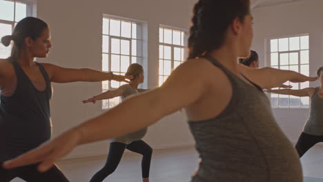 yoga class of healthy pregnant women practicing warrior pose enjoying group physical fitness workout with instructor in studio at sunrise