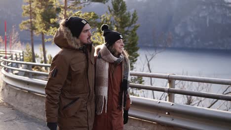 tourists in winter coats and black hats walking in slow motion on a long road through the countryside towards the snowy mountain peaks and lake on the background. smiling caucasian couple exploring norway nature