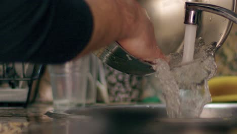 Cerca-De-Un-Hombre-Limpiando-Una-Olla-Y-Vertiendo-Agua-Durante-Las-Tareas-Domésticas-En-Casa-En-La-Cocina-En-Cámara-Súper-Lenta-Por-La-Noche-Tiro-Medio