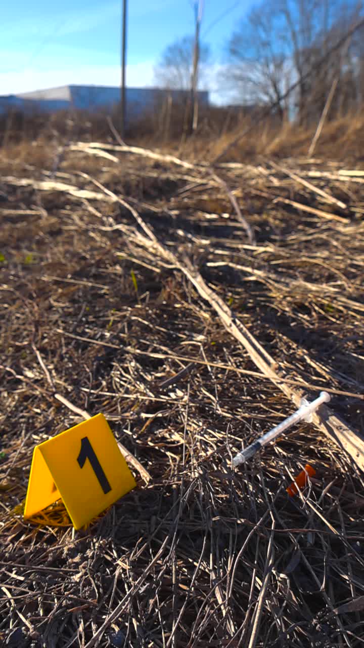 Primo piano di una siringa usata con il suo cappuccio arancione adagiata nell'erba secca accanto al numero uno del marcatore di prova della polizia giallo su una scena del crimine all'aperto in una giornata di sole