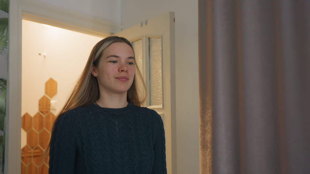 Lady wearing black sweater and simple black necklace walks through white painted door into softly lit interior with textured glass panels and patterned wallpaper, captured in casual indoor setting