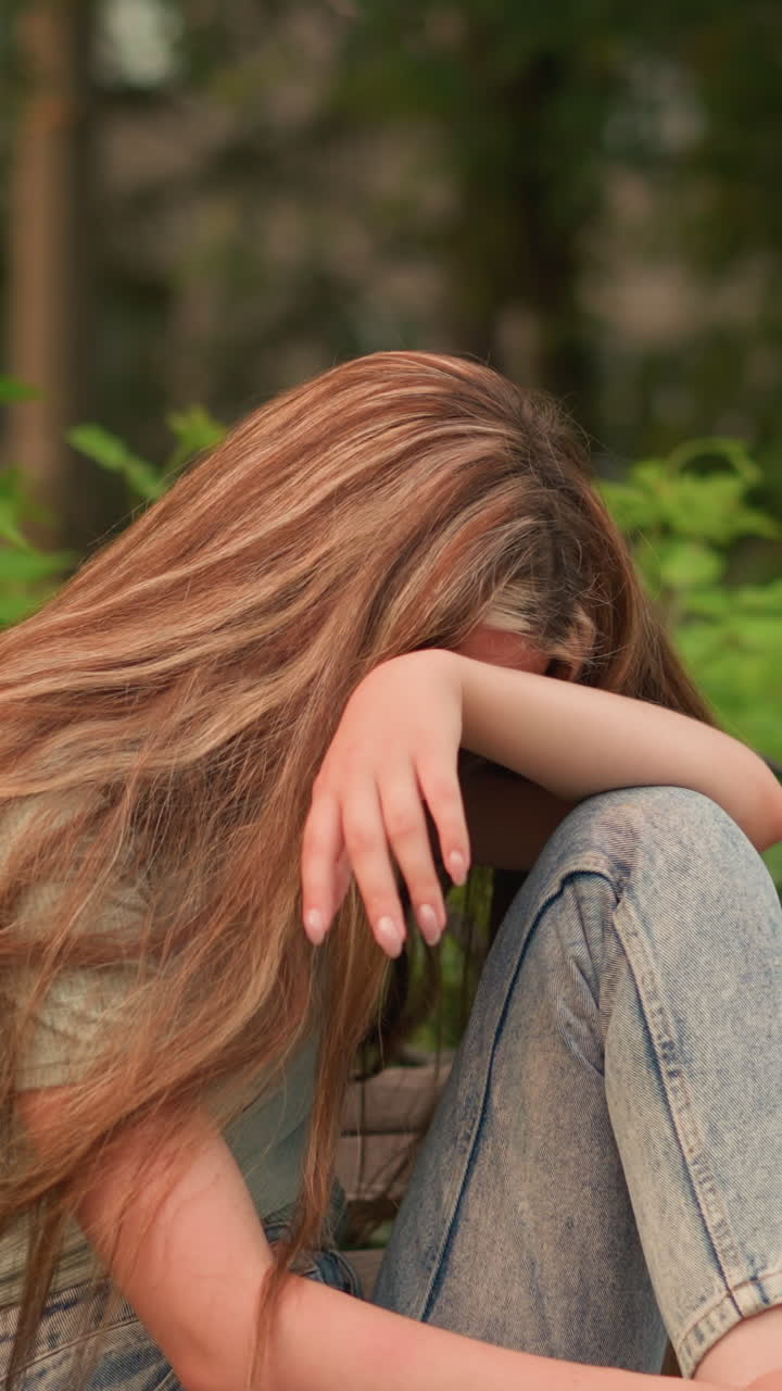 청바지와  스니커를 입은 젊은 여성은 공원의 나무 벤치에 앉아서 머리를 팔 위에 놓고 반사적이고 우울한 자세로 은 손과 어깨 위에 흐르는 긴 머리카락을 가지고 있습니다.