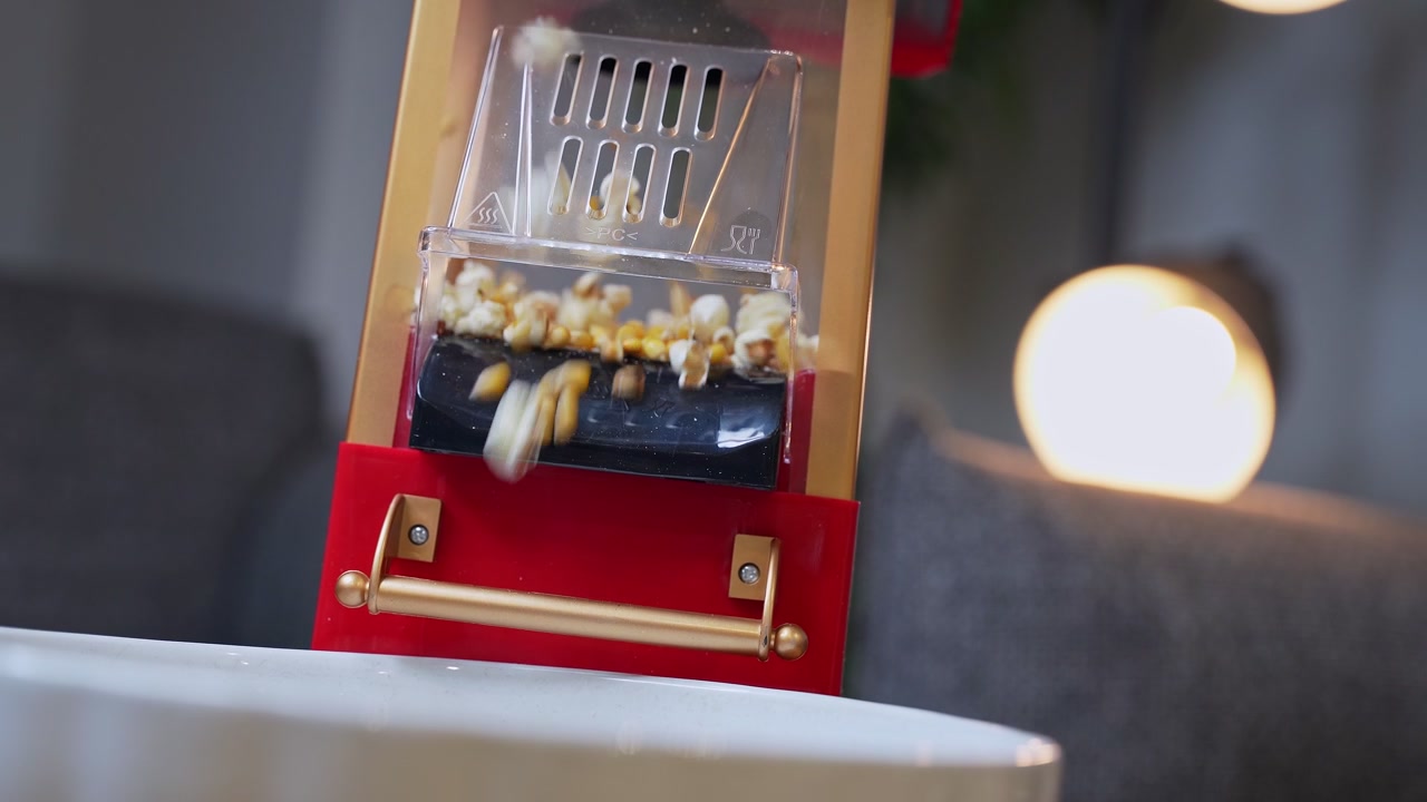 a vintage red and gold popcorn machine popping fresh corn kernels into a white bowl in a cozy home interior with warm bokeh lighting. Perfect for cinema and snack-themed content.