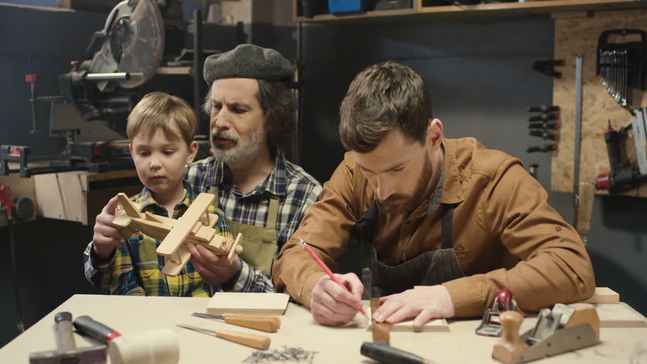 carpintero anciano sentado en el escritorio en el taller con su hijo y su lindo nieto