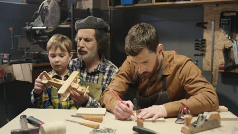 carpintero anciano sentado en el escritorio en el taller con su hijo y su lindo nieto