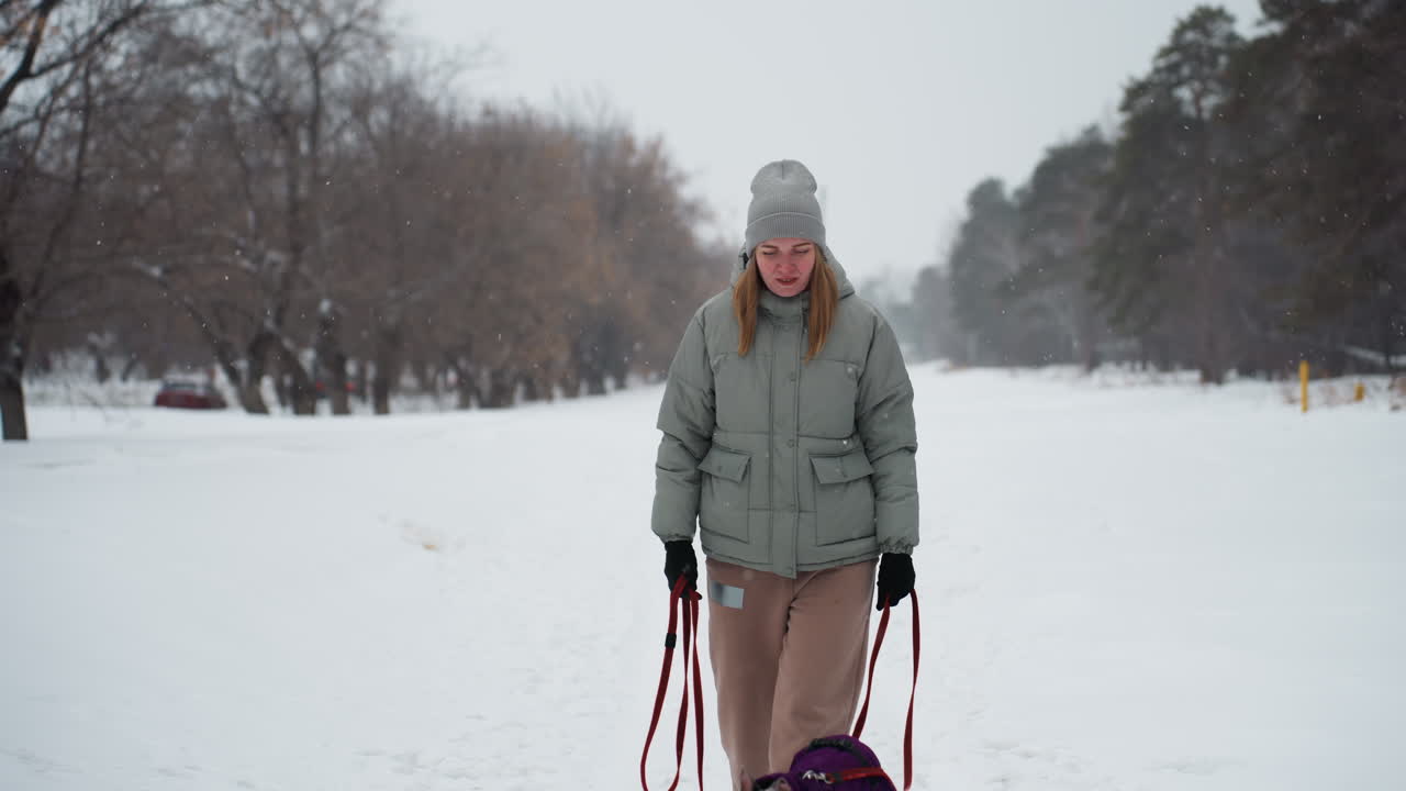 Young woman dressed in winter clothing walks snowy path holding red leash, partially visible dog at bottom, overcast sky above, surrounded by leafless trees and forest edge during gentle snowfall