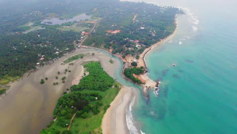 Aerial,-tilt,-drone-shot-of-waves-hitting-a-paradise-coast,-revealing-Trincomalee-town,-on-a-cloudy-day,-in-Gokanna,-in-the-Eastern-Province-of-Sri-Lanka