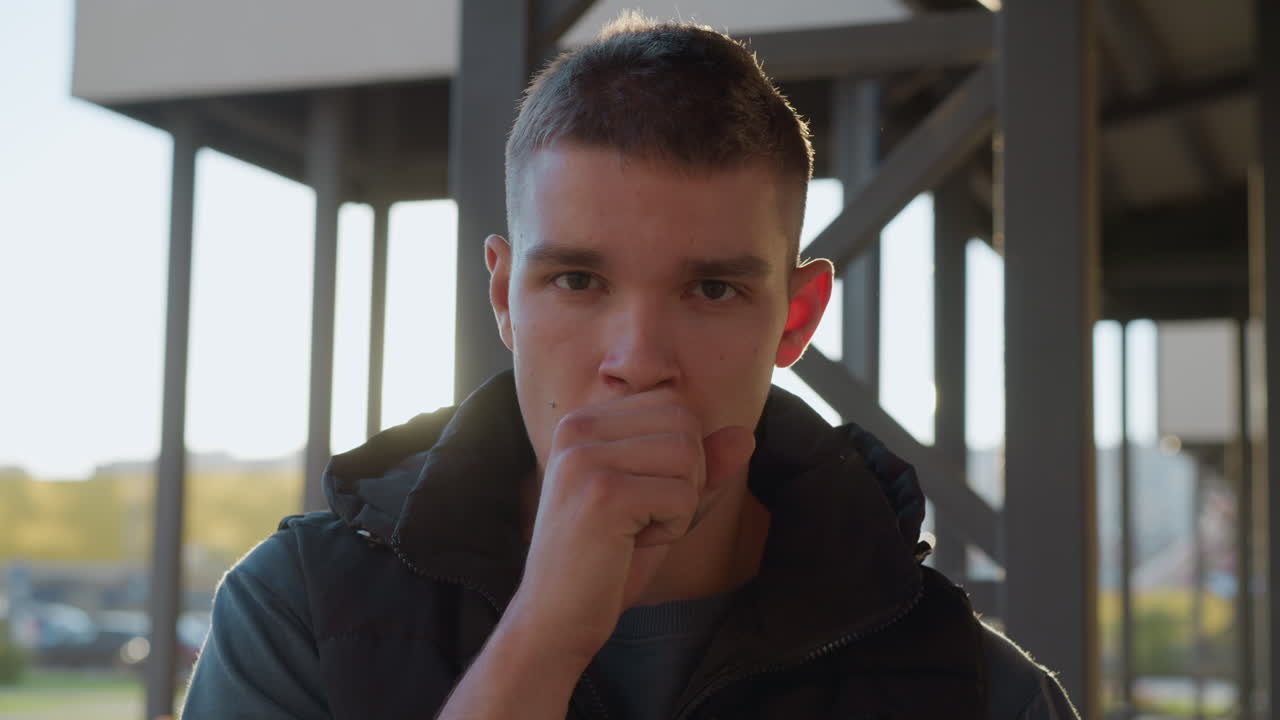 portrait view of young man outdoors coughing into fist while standing in soft backlight under urban structure showing discomfort or reaction to irritation in throat during cool morning air