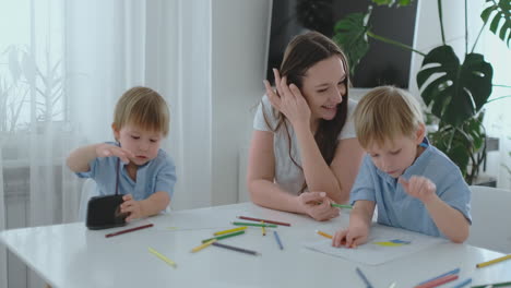 Mamá-Sentada-A-La-Mesa-Con-Dos-Hijos-Niño-Afilando-Un-Lápiz-De-Color-En-Un-Sacapuntas-Sentado-En-La-Mesa-De-La-Cocina.-El-Segundo-Niño-Hace-Un-Dibujo-Con-Lápices