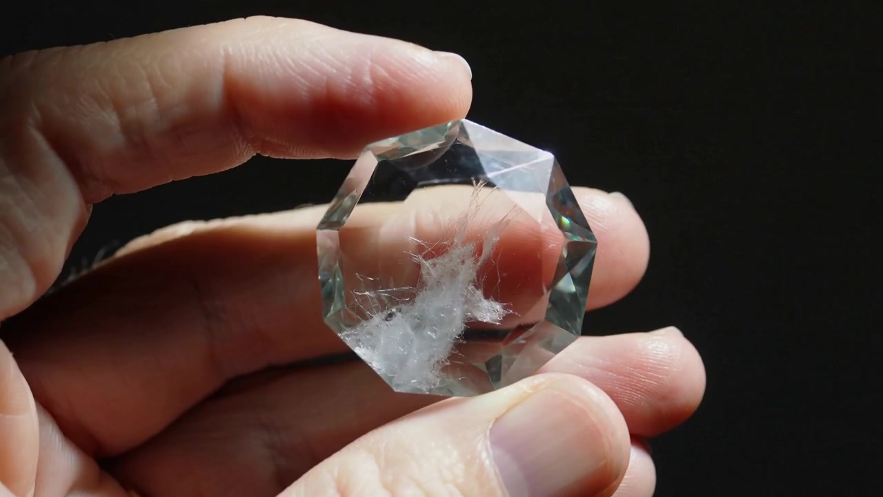 Una mano adulta prende e ruota un cristallo sfaccettato in studio, esaminando un'inclusione bianca e un anello d'oro.