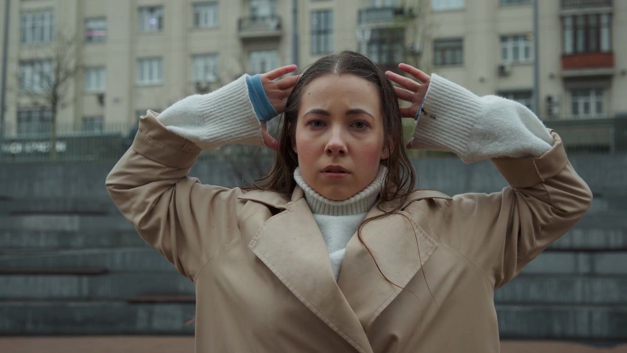 Primo piano di una giovane donna con lunghi capelli che indossa guanti di lana bianchi e un cappotto beige, che muove le mani vicino al viso in un ambiente urbano esterno. La donna interagisce sottilmente alzando le braccia o aggiustando.