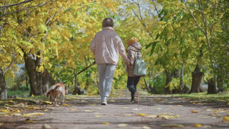 woman holding dog on leash as it tries to break free while daughter walks beside under autumn canopy, leaves scattered on path, playful struggle and family outdoor stroll in golden forest light