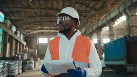 Un-Hombre-De-Piel-Negra-Con-Casco-Protector-Y-Uniforme-Blanco-Con-Chaleco-Naranja-Camina-Entre-Los-Estantes-De-Residuos-Reciclados-Y-Clasificados.