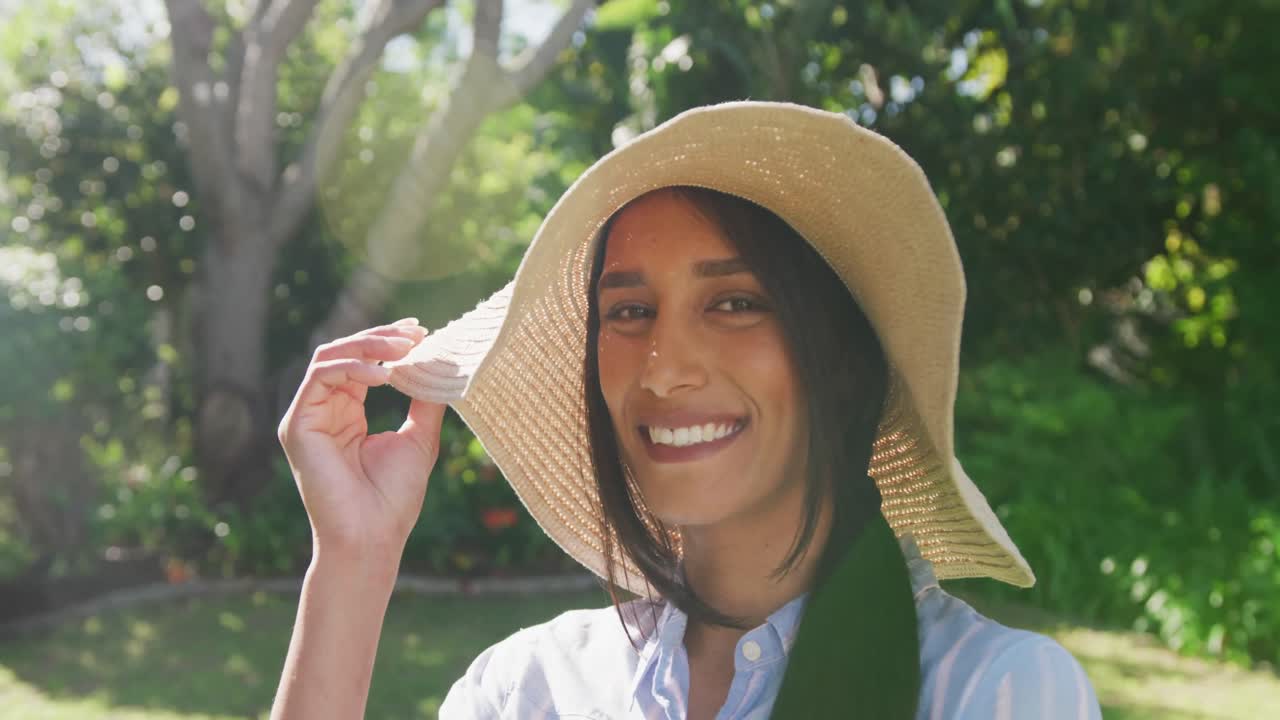 retrato de video de una mujer biracial feliz con sombrero de sol en un jardín soleado sonriendo a la cámara